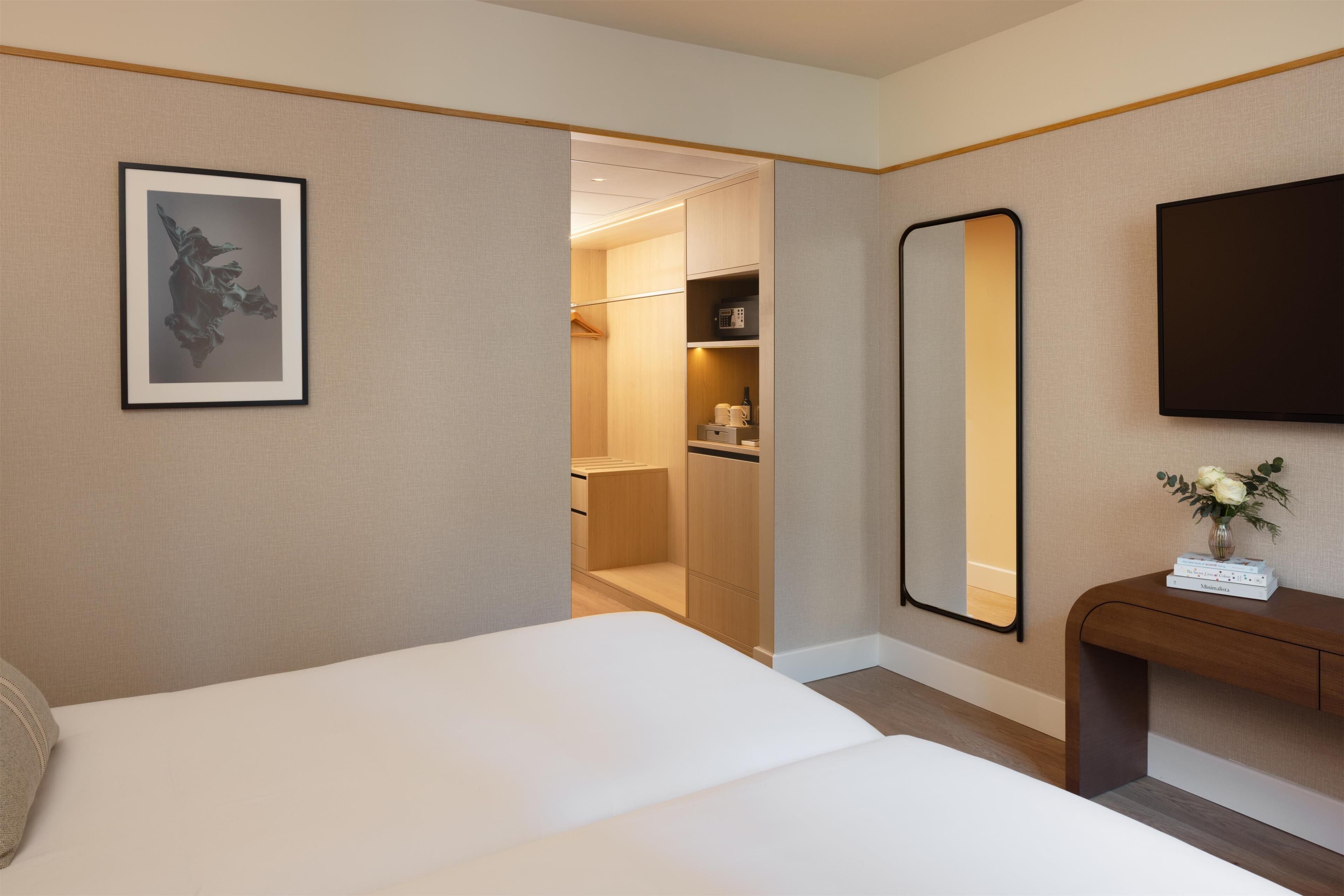 A hotel room with a bed, a bedside table with a vase, a large mirror, and a wardrobe with a hanging rack.