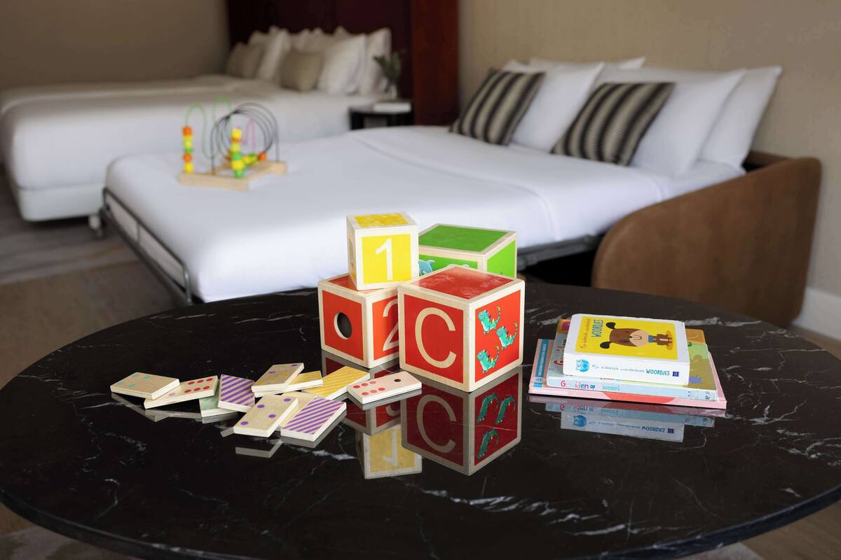 Una habitación de hotel con una cama, una mesita de noche con un juguete y una mesa con libros y dados.
