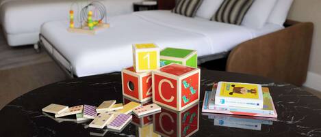 Una habitación de hotel con una cama, una mesita de noche con un juguete y una mesa con libros y dados.