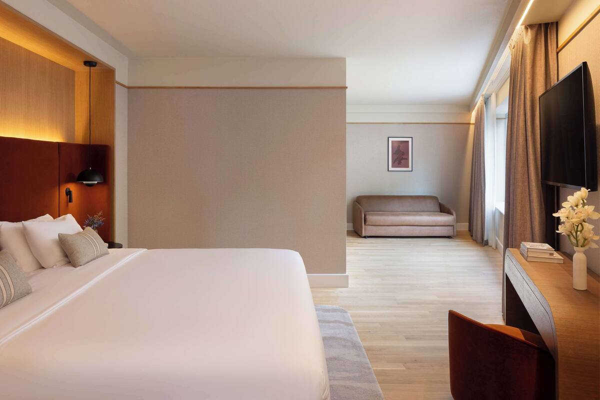 Una habitación de hotel con una cama grande, un cabecero de madera, un televisor de pantalla plana, un sofá y un escritorio con un jarrón de flores.