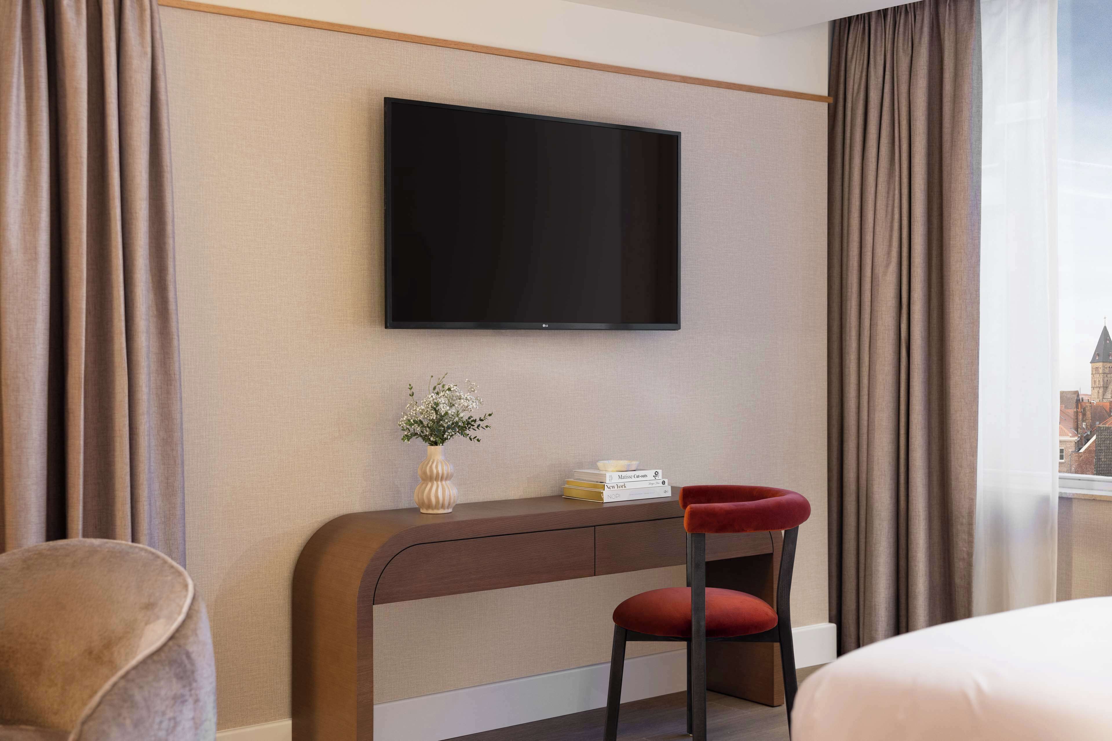 A hotel room with a flat-screen TV mounted on the wall, a desk with a chair, a vase with flowers, and a view of a cityscape through a window.