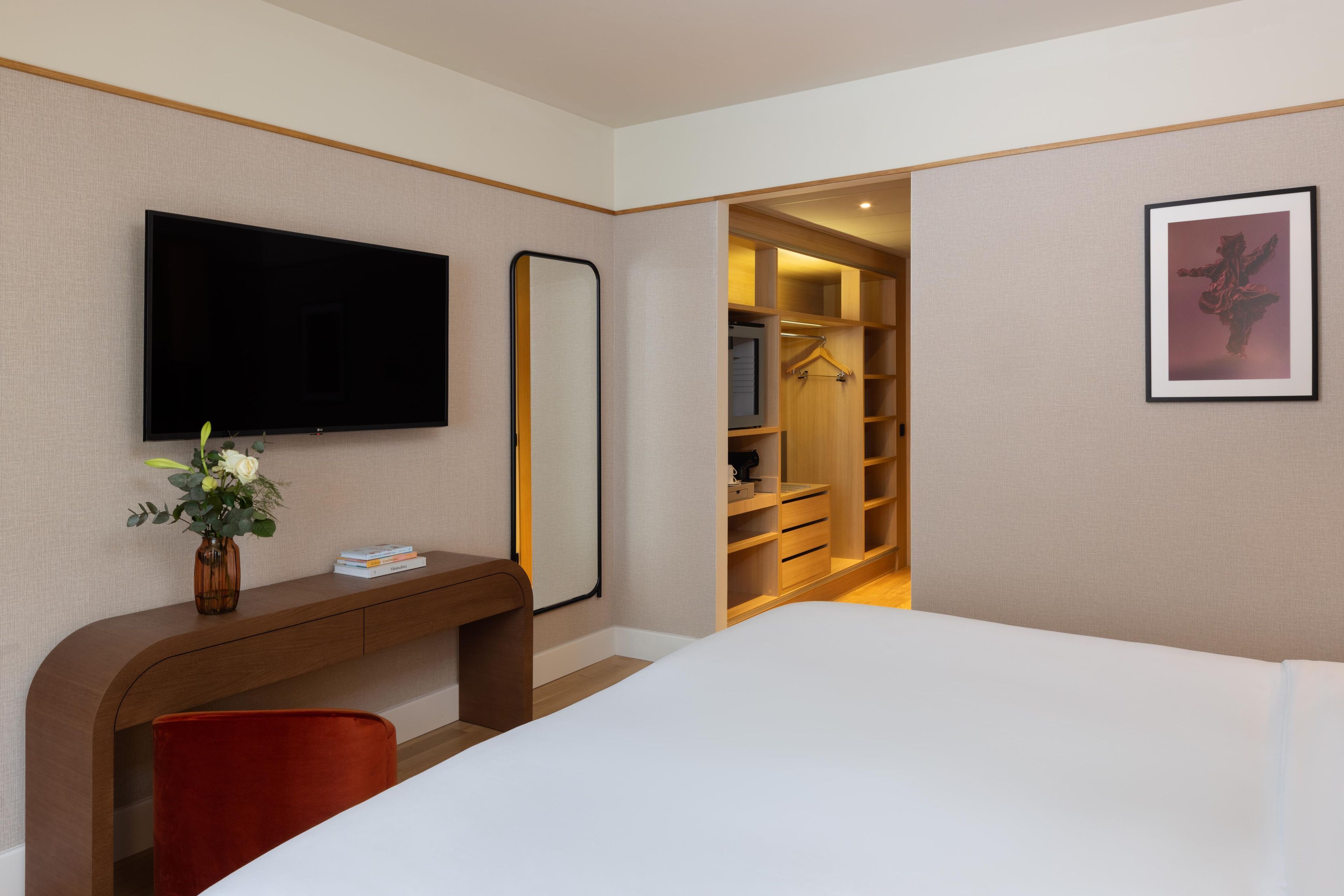 A hotel room with a bed, a desk with a vase of flowers, a television, a wardrobe, and a framed picture on the wall.