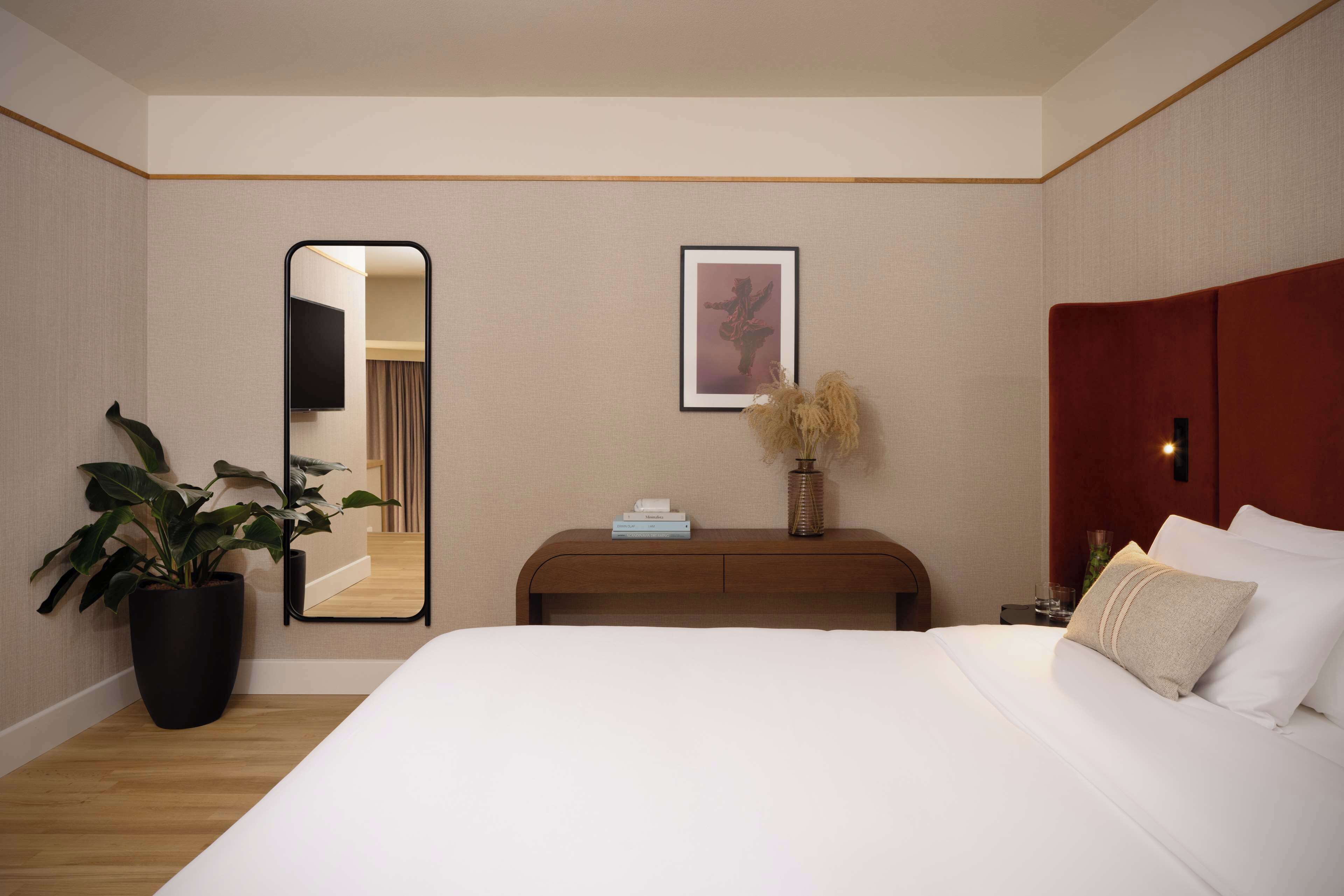 A hotel room with a large bed, a bedside table with a vase, a mirror, and a framed picture on the wall.