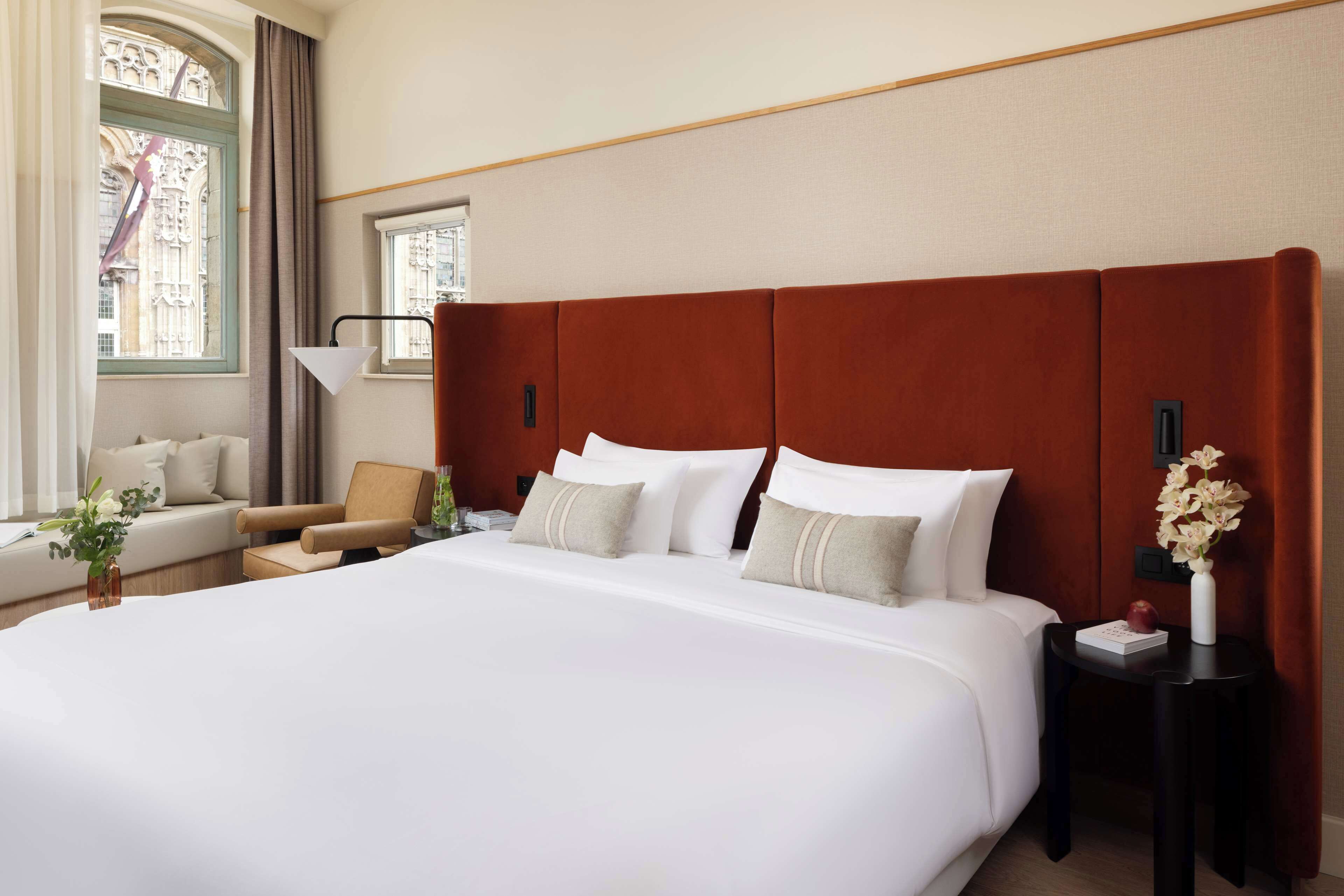 Habitación de hotel con una cama grande, un sofá, una silla, una mesita de noche y una ventana con vistas a un edificio.