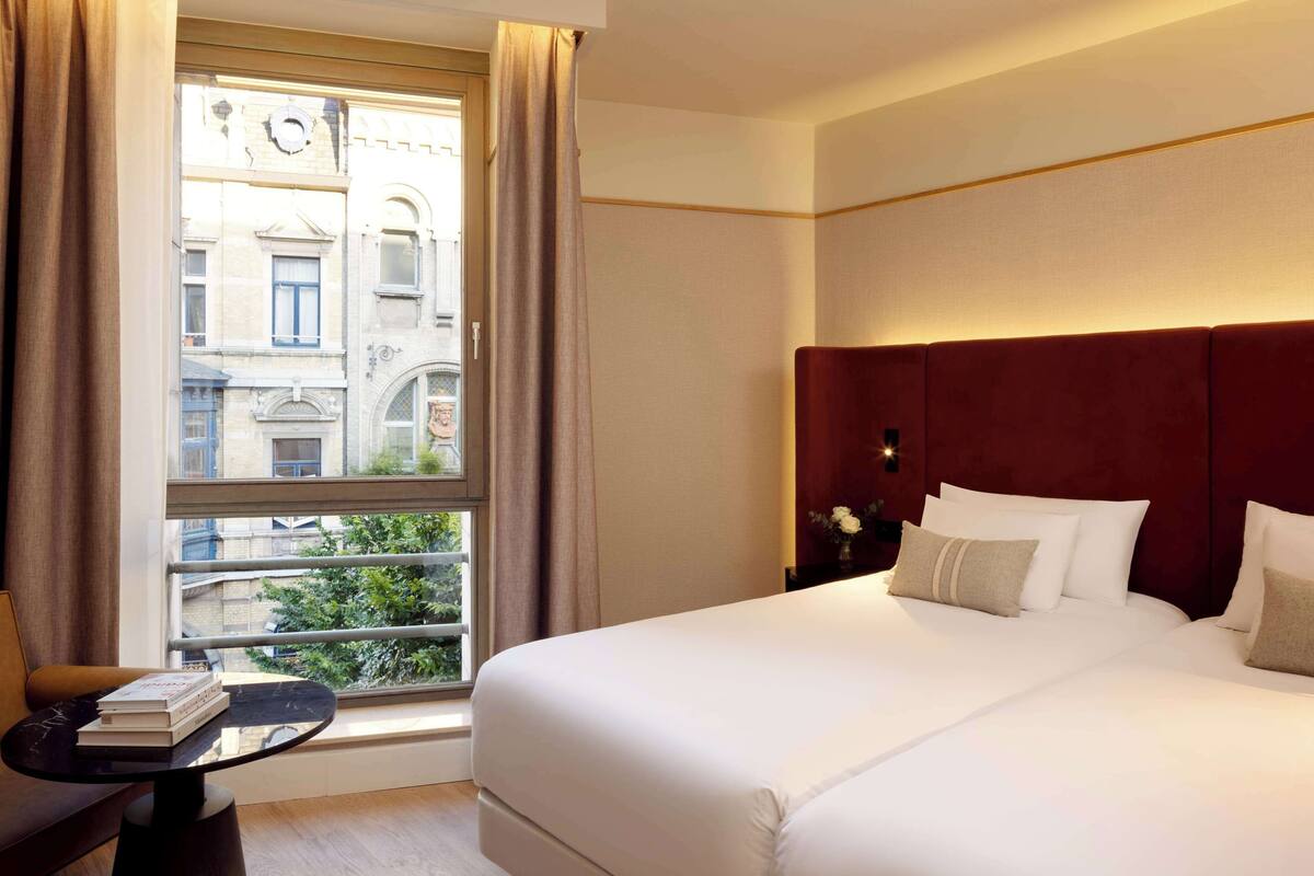 Habitación de hotel con una cama grande, una mesita con libros, una ventana con cortinas y vistas a edificios.