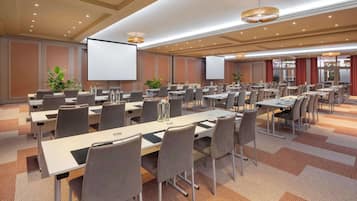 A conference room with multiple tables and chairs, equipped with projectors and screens, and decorated with plants.