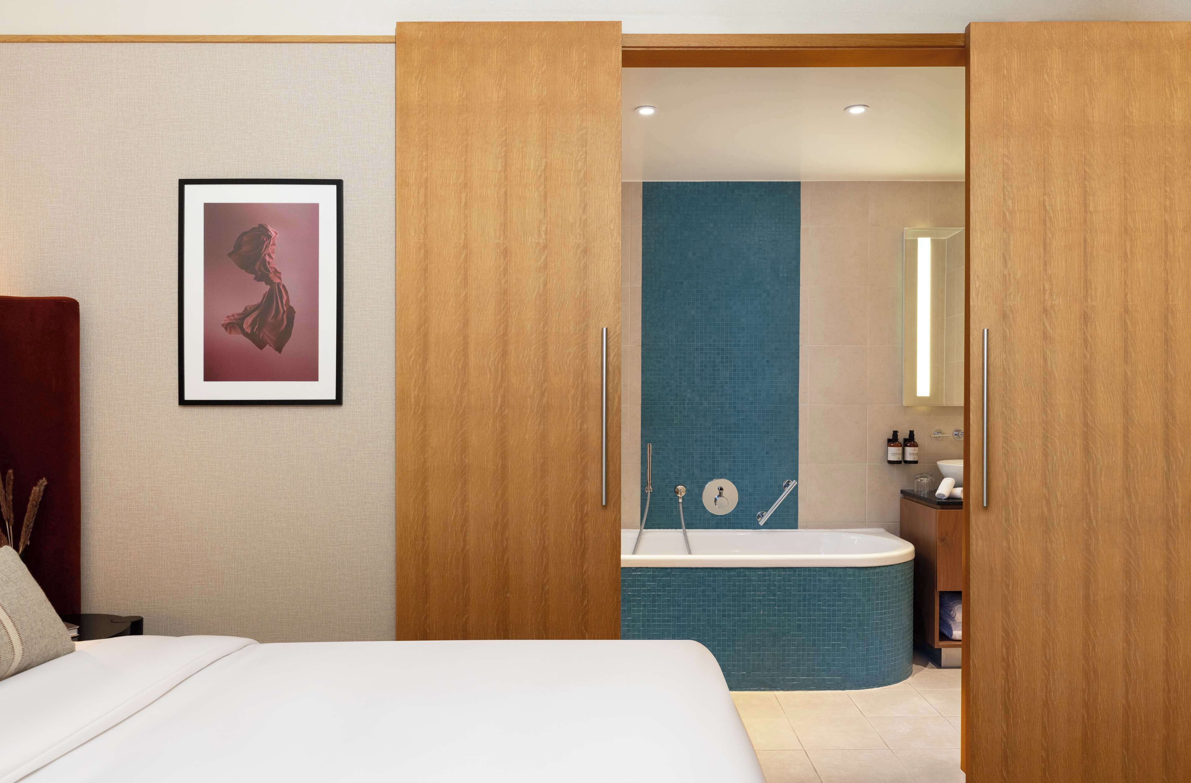 Una habitación de hotel con cama, un cuadro enmarcado en la pared, un armario de madera y un baño con bañera y lavamanos.