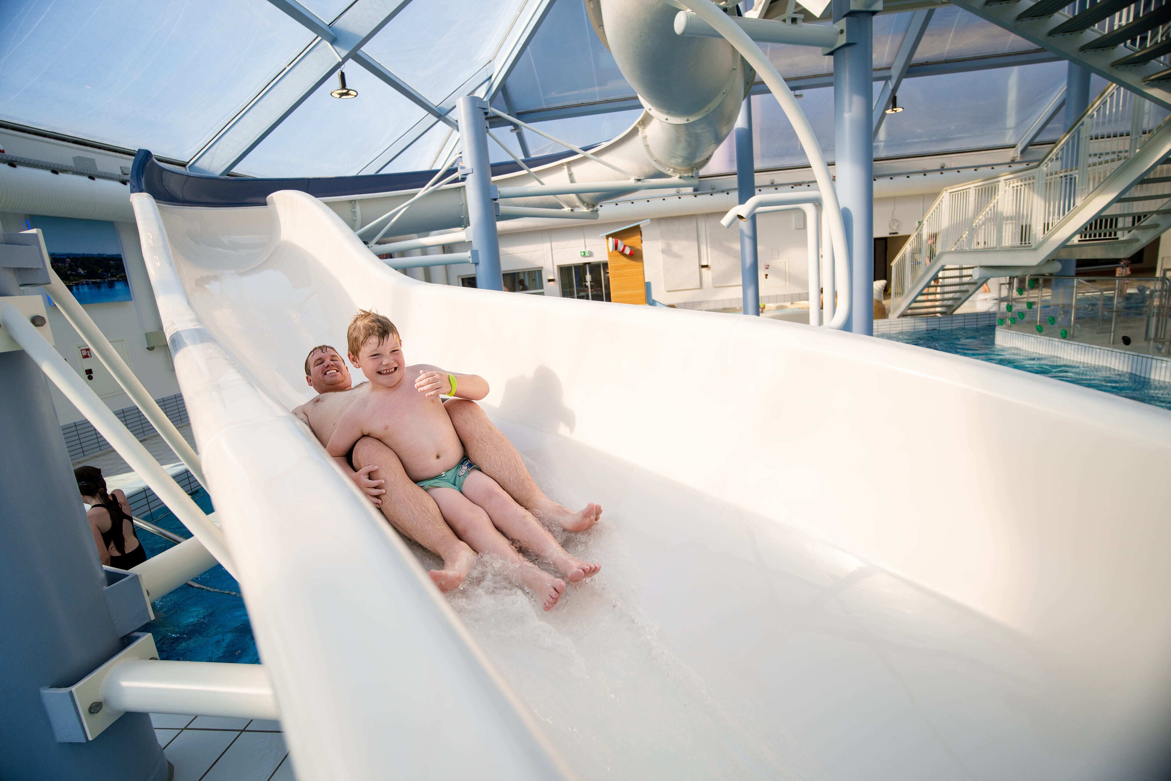 Un tobogán de agua en un crucero con techo transparente y una zona de piscina visible al fondo.