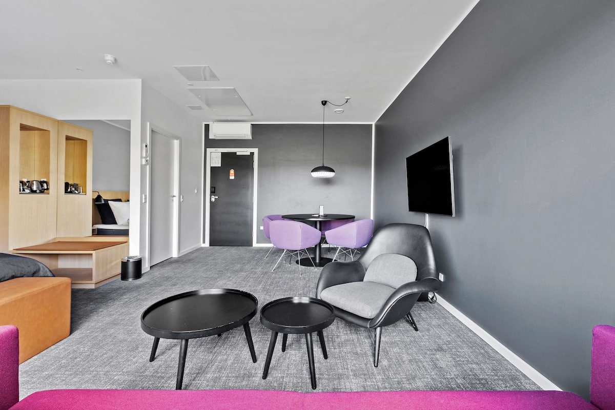 Una habitación de hotel moderna con alfombra gris, un sofá, dos mesas redondas, una silla y un televisor montado en la pared.