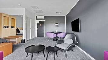 A modern hotel room with a grey carpet, a sofa, two round tables, a chair, and a TV mounted on the wall.