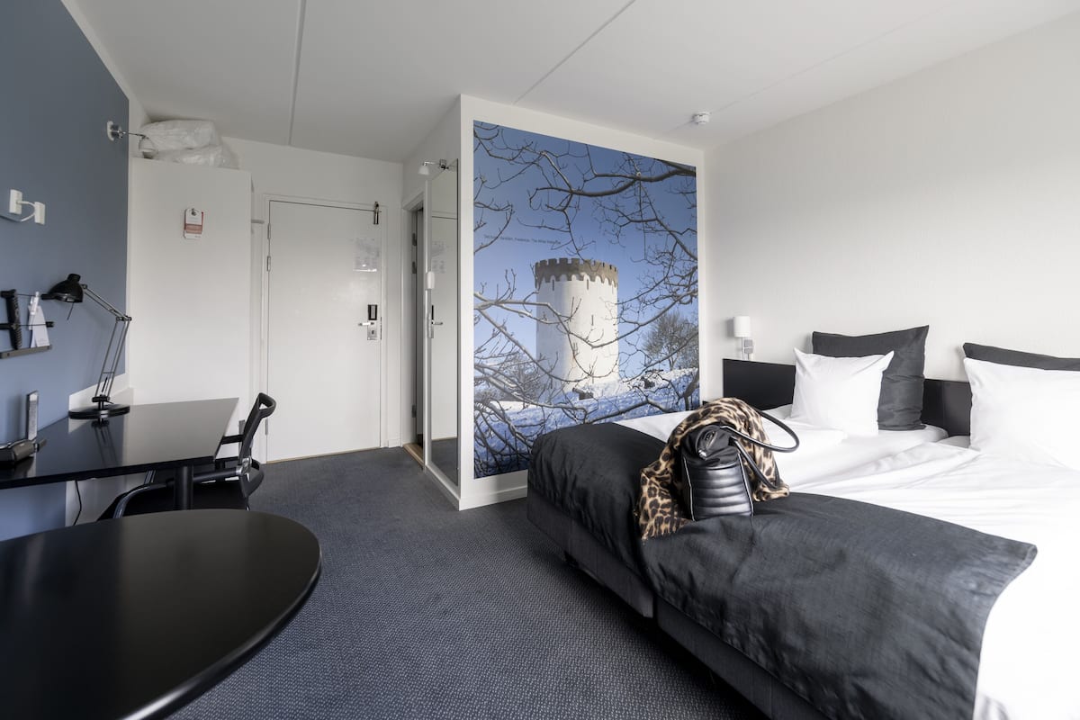 Habitación de hotel con cama, escritorio y silla. Hay un cuadro en la pared y una ventana con vista a una torre.