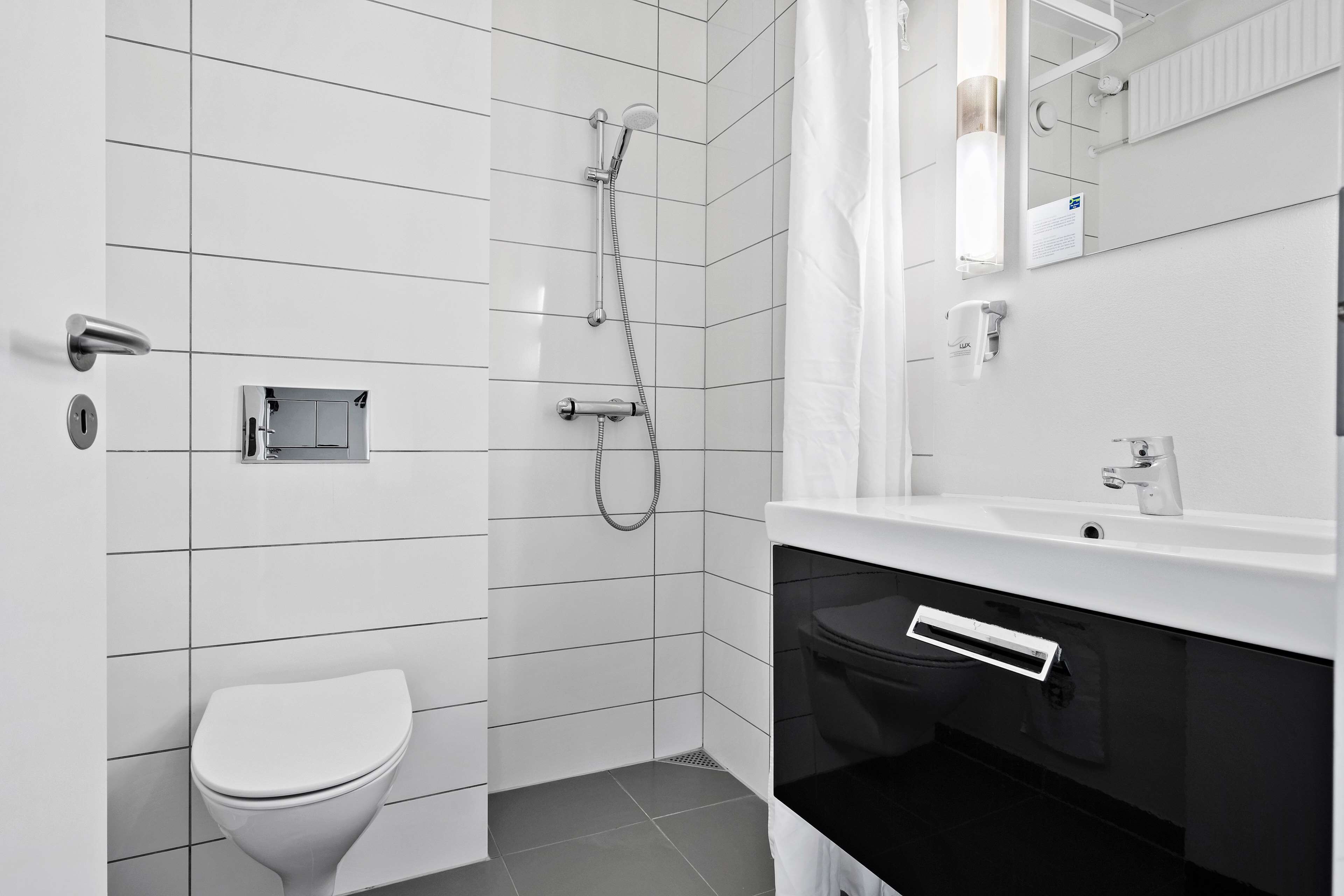 Et moderne badeværelse med hvide fliser, toilet, brusebad med håndbruser, vask med spejl og et håndklædetørrestativ.