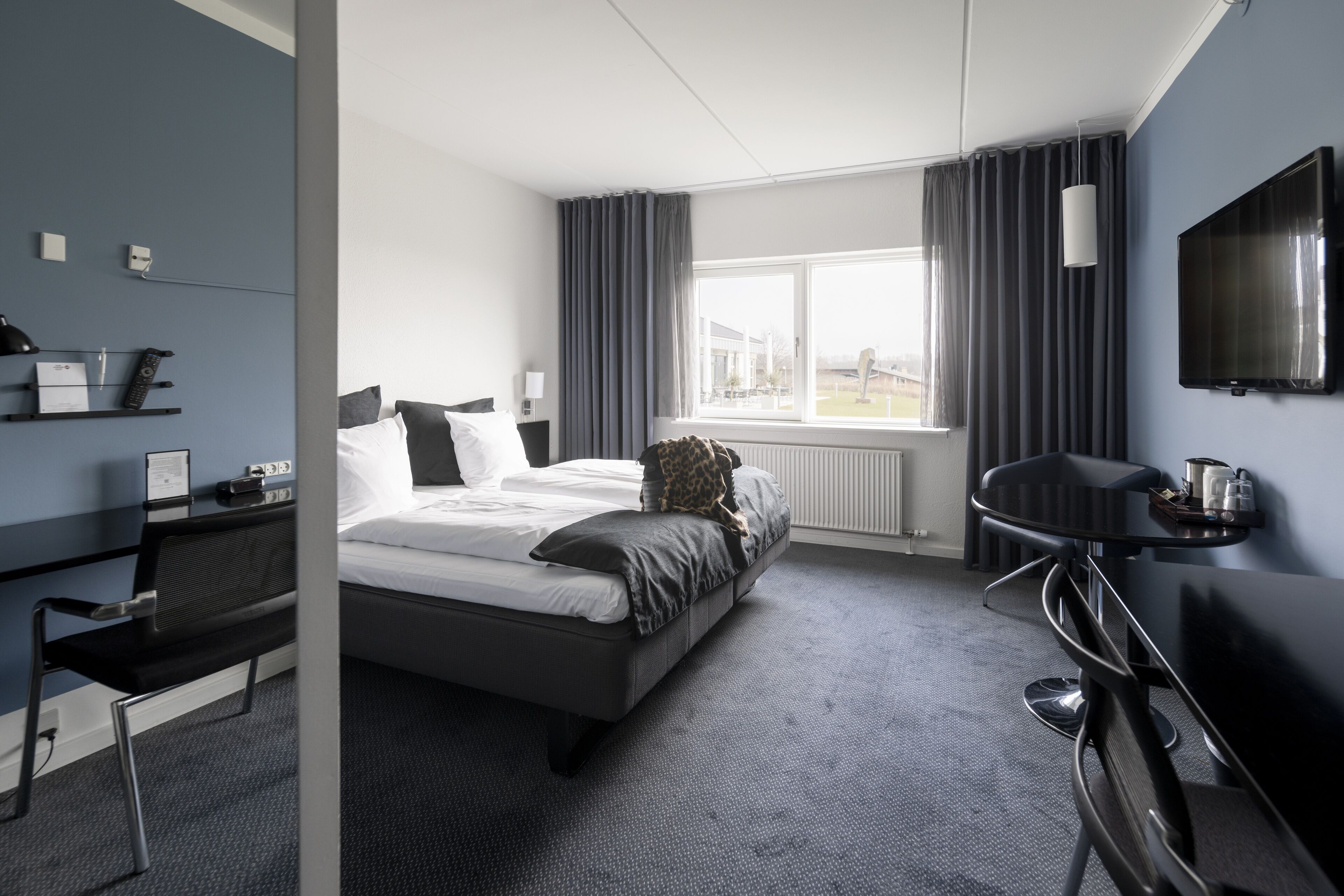 Una habitación de hotel moderna con cama, escritorio, silla, televisión y ventana con cortinas.