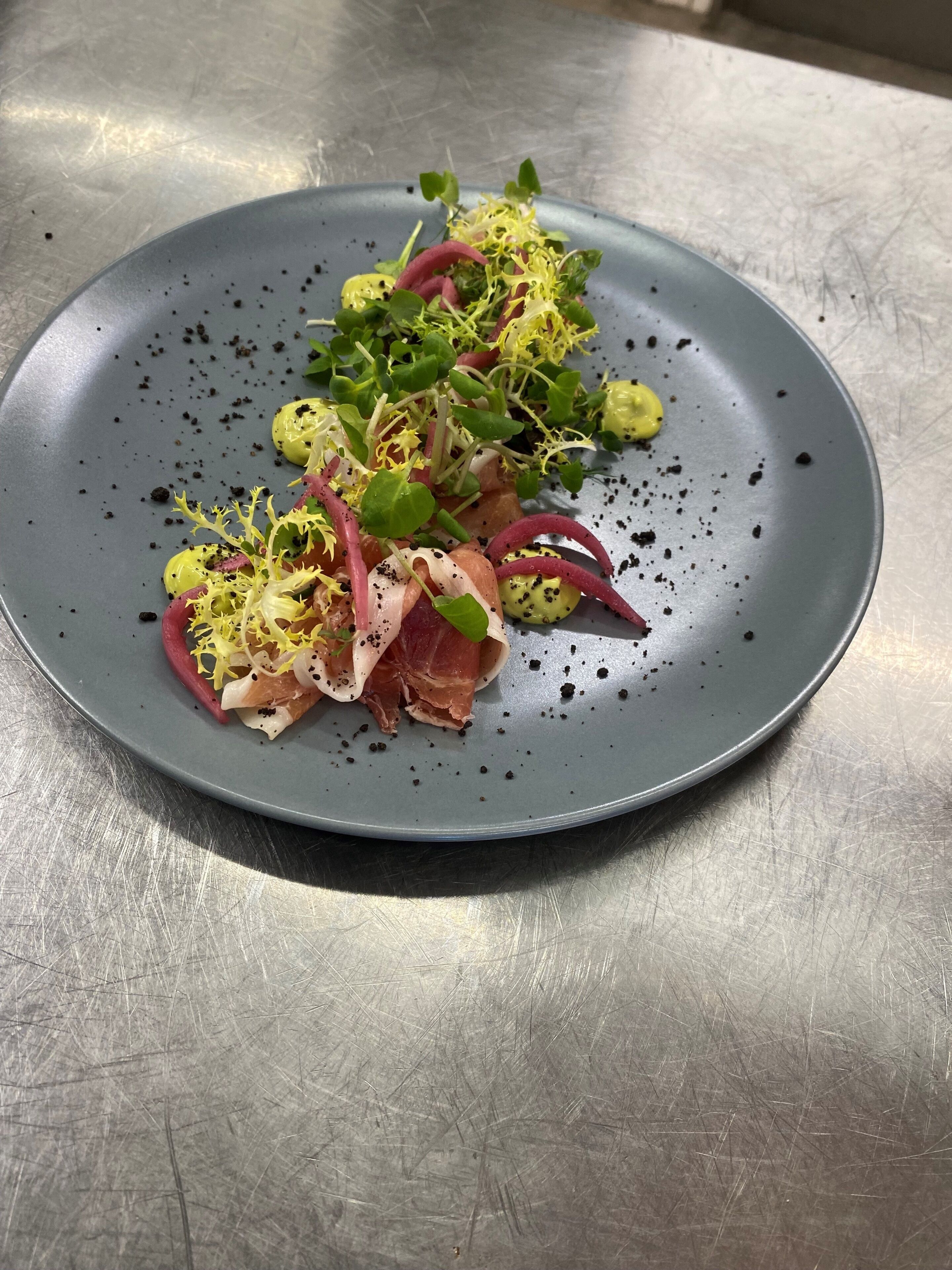 A plate with a variety of food items including greens, sprouts, and slices of meat, served on a grey plate.