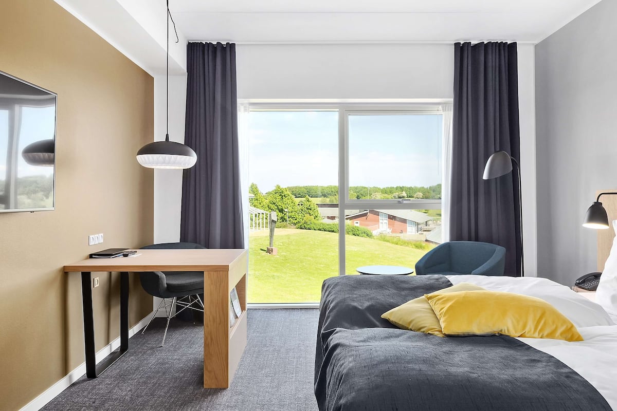 Habitación de hotel con cama, escritorio, silla y un amplio ventanal con vistas al exterior.
