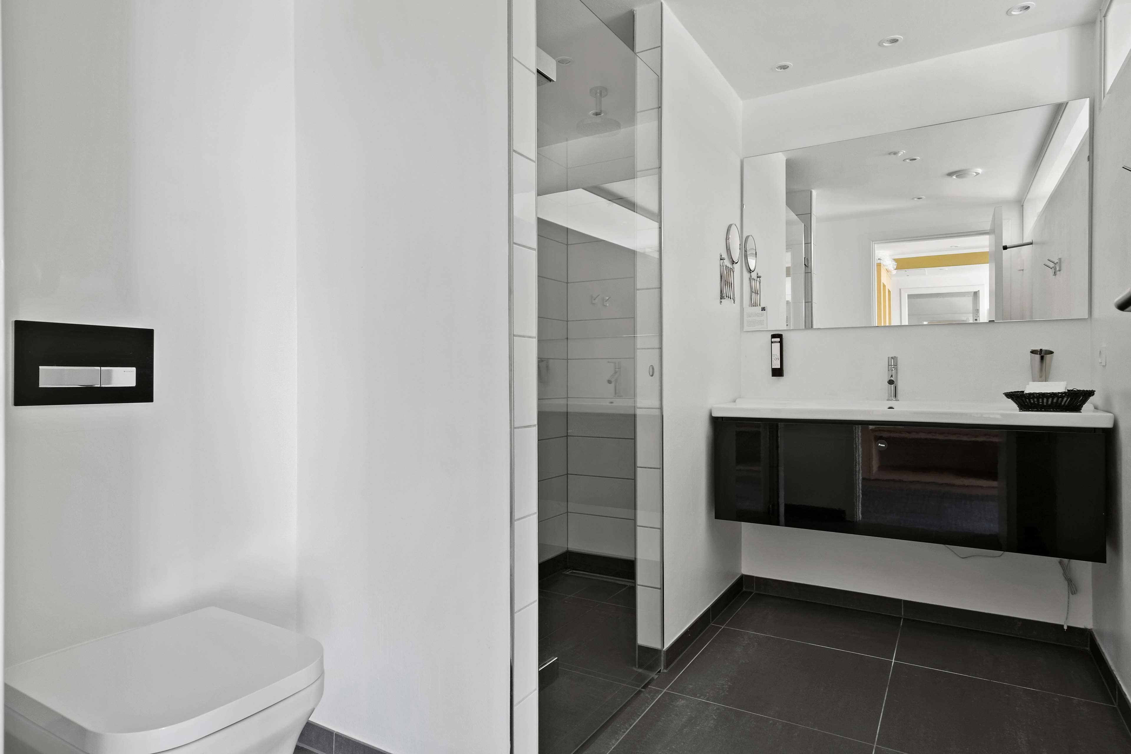Een moderne badkamer met een wit toilet, een douchecabine van glas, een wastafel met een zwarte wasbak en een grote spiegel.