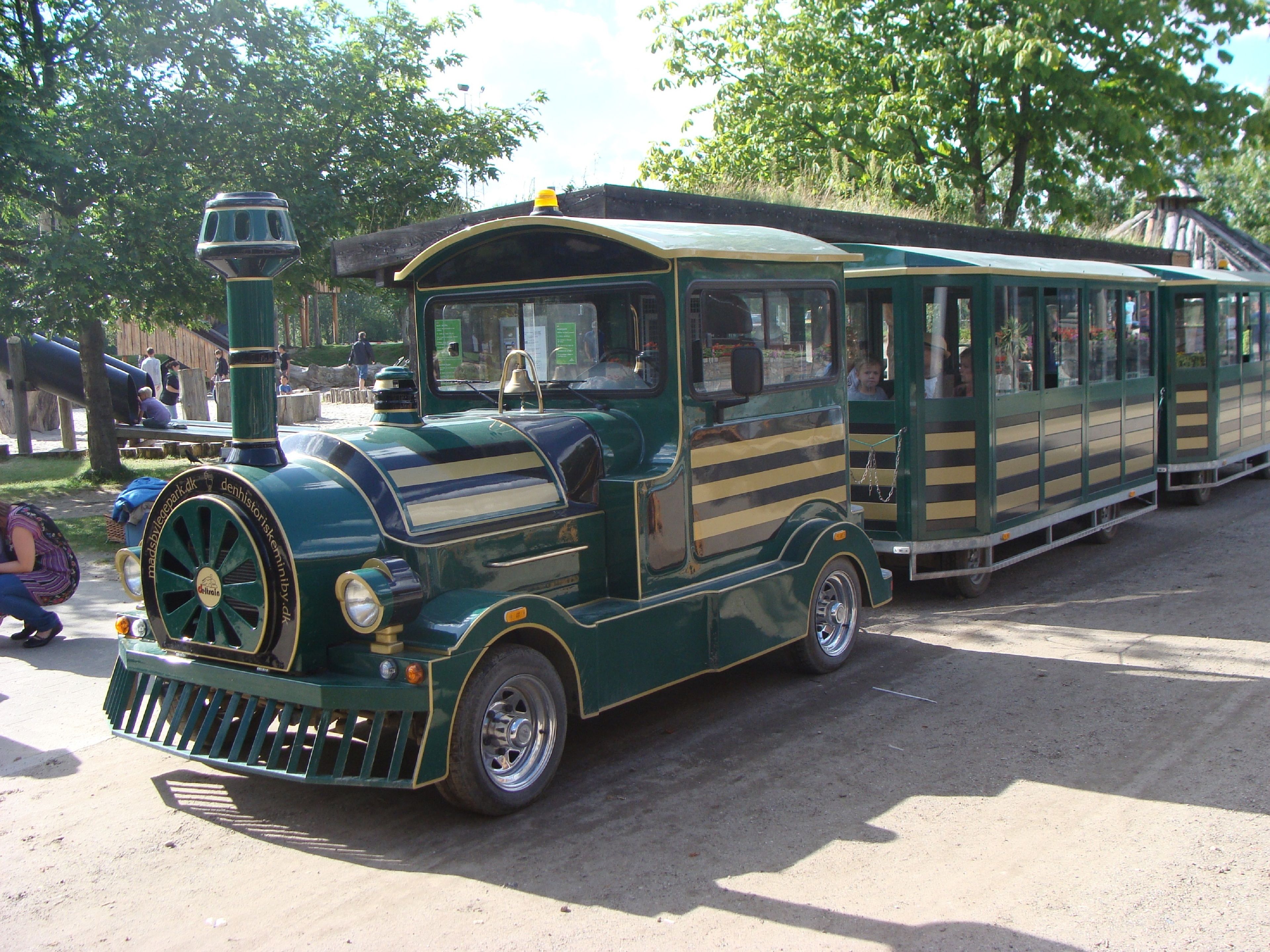 Un tren verde antiguo con una campana en la parte delantera y una cabina pequeña, estacionado en un sendero de tierra.