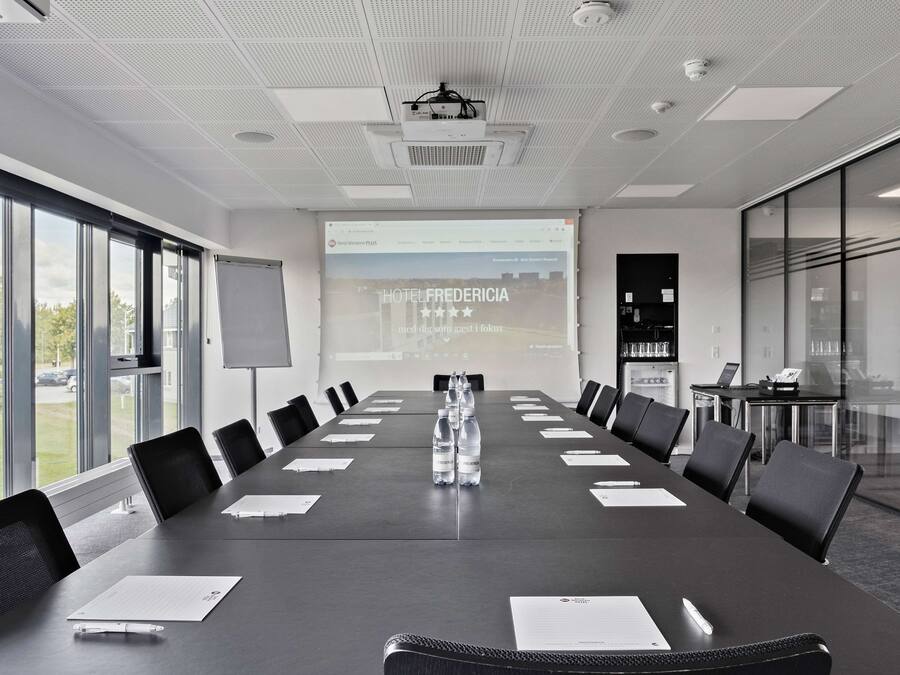 Ein Konferenzraum mit einem langen Tisch, Stühlen, einer Projektionsleinwand und einer Präsentation über das Hotel Fredericia.