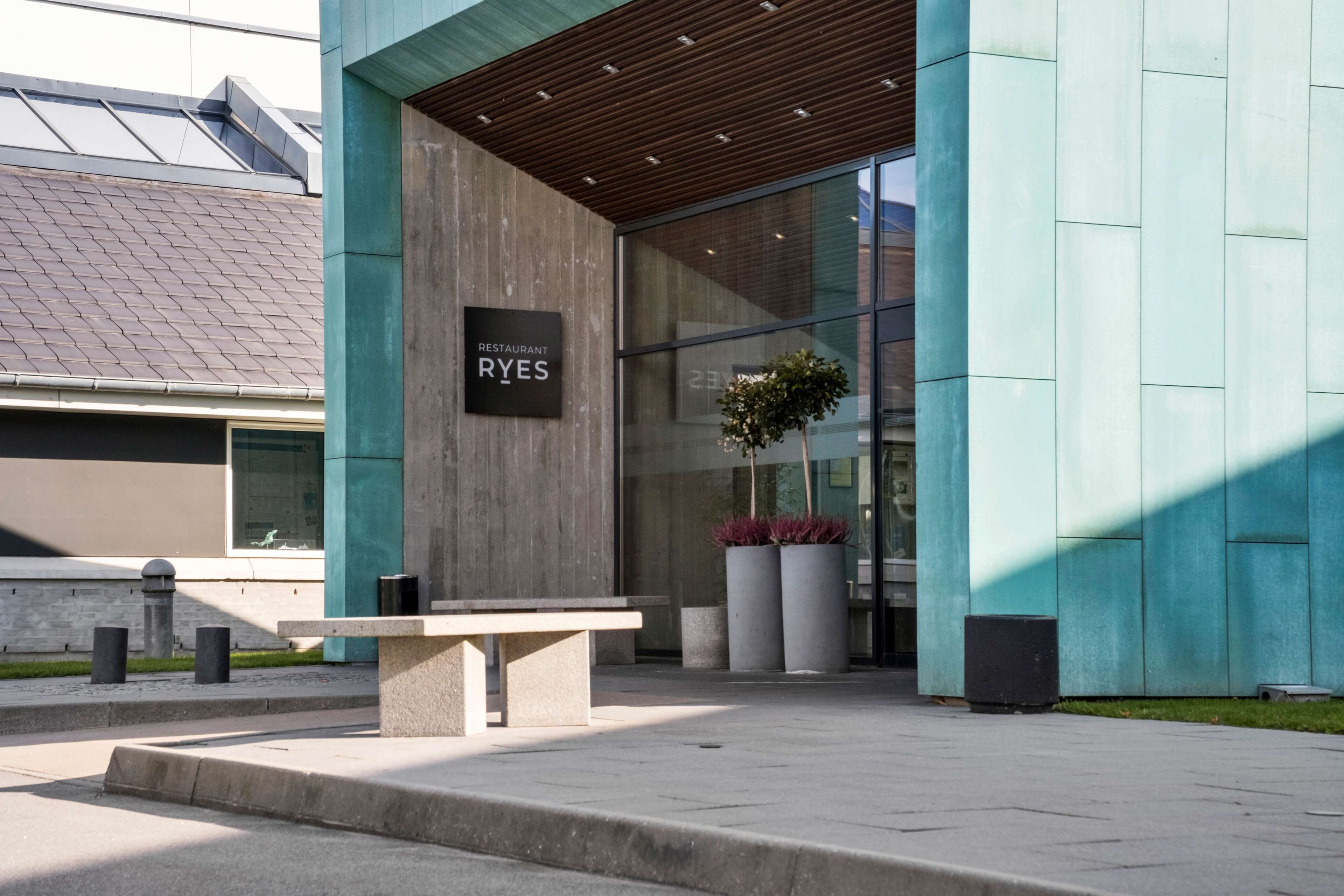 Une entrée moderne avec une enseigne indiquant « Restaurant RYES », un banc et de grands bacs à plantes.