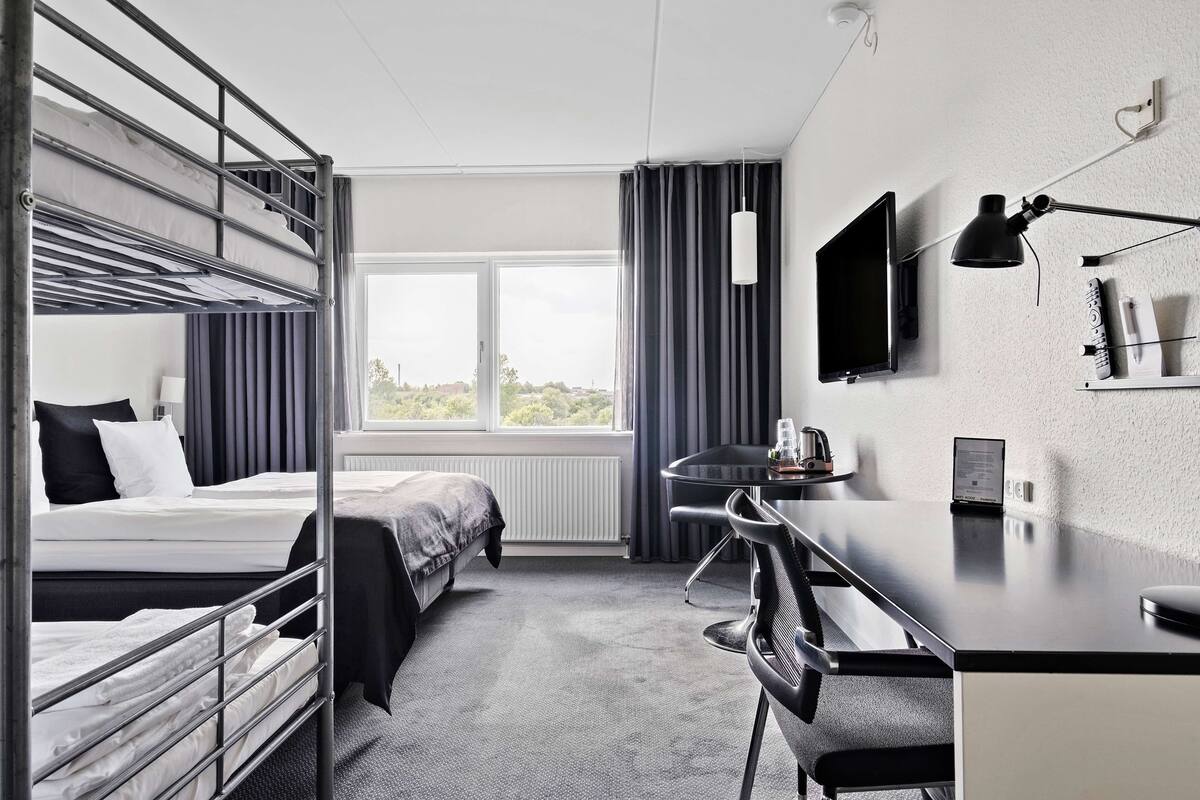 Una habitación de hotel con literas, un escritorio con silla, un televisor y una ventana con cortinas.