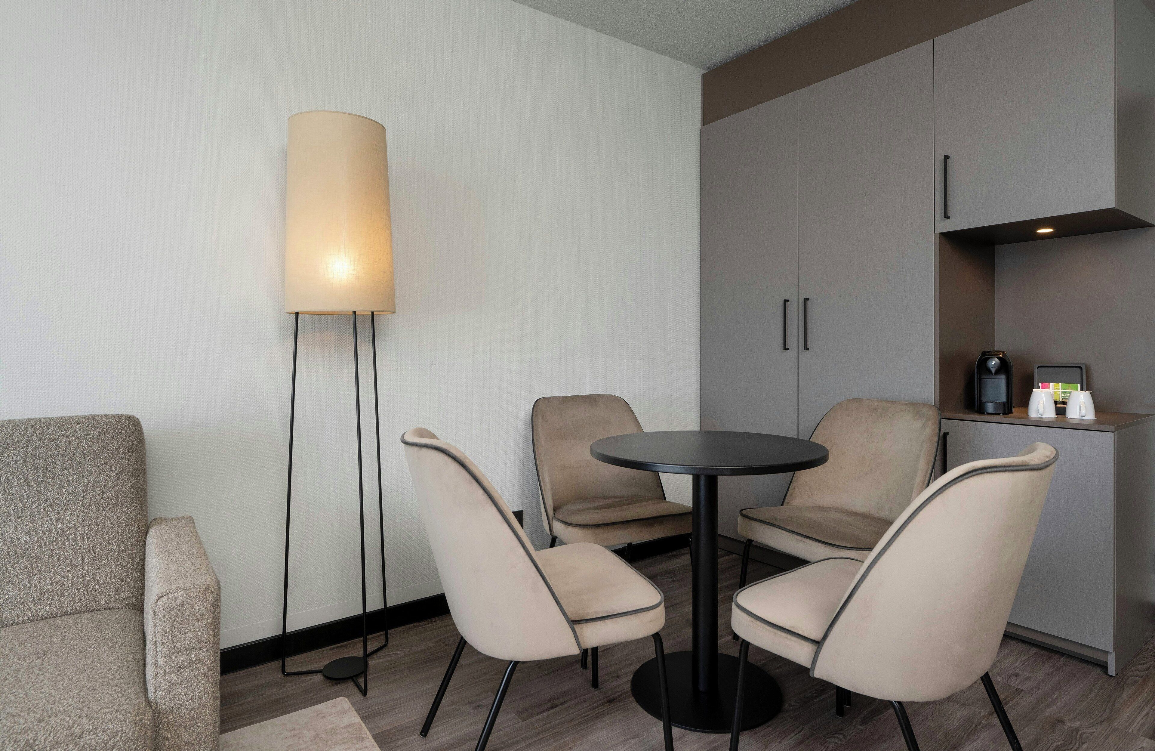 Habitació moderna amb taula rodona, dues cadires, làmpada de peu i zona de cuina equipada.