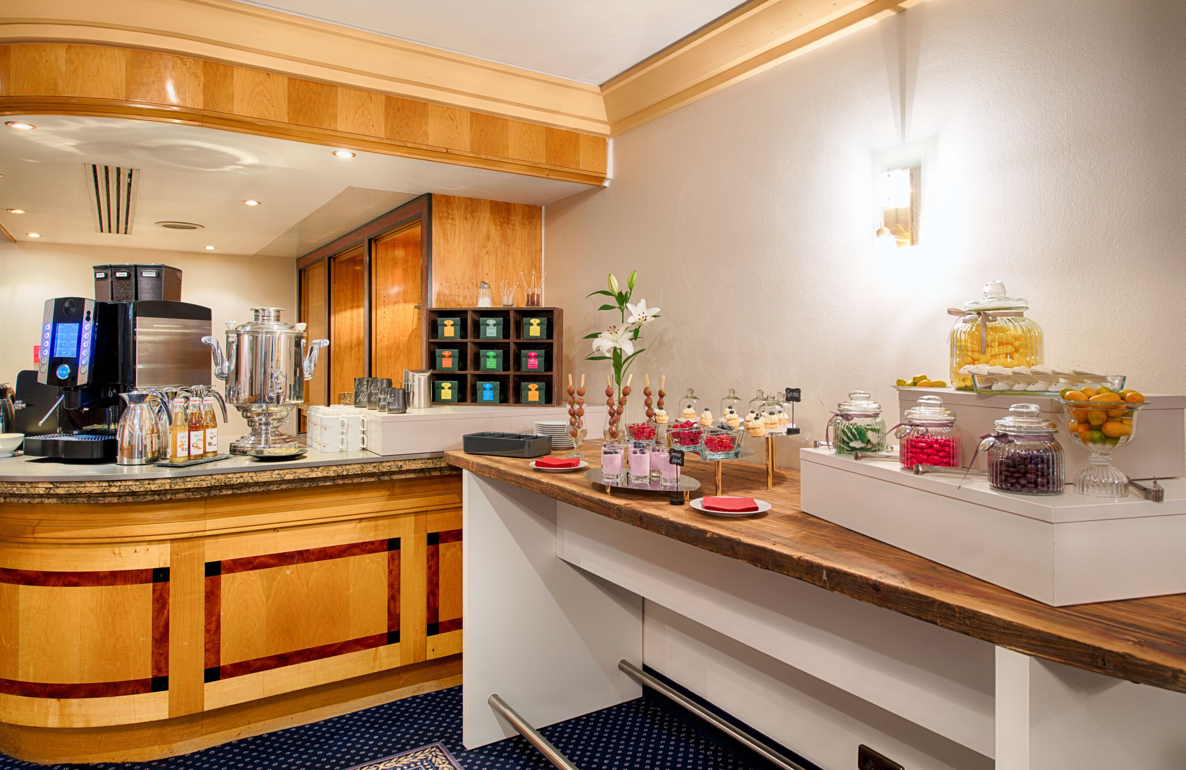 A hotel breakfast area with a coffee machine, tea and coffee dispensers, and a variety of food and drink options.