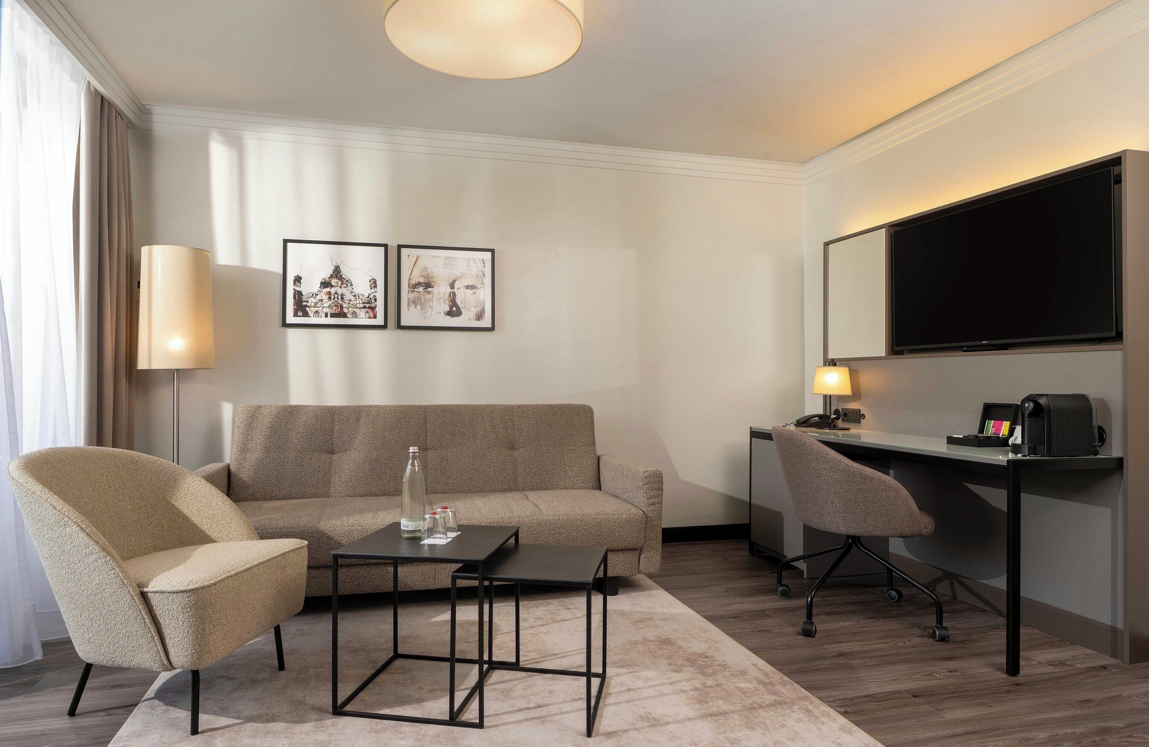A modern hotel room with a sofa, armchair, desk, and a flat-screen TV.