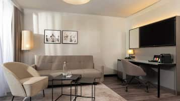 A modern hotel room with a sofa, armchair, desk, and a flat-screen TV.