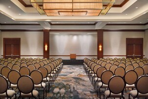 Una sala conferenze con file di sedie rivolte verso un palco, pannelli in legno sulle pareti e una moquette decorata.