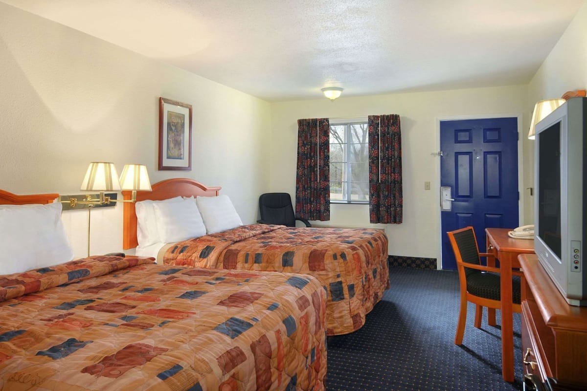 Une chambre d’hôtel avec deux lits, un bureau avec une chaise, une télévision et une fenêtre avec des rideaux.