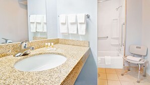 Une salle de bain avec un lavabo sur un comptoir en granit, un miroir et un espace douche équipé d’une barre d’appui et d’une chaise blanche.