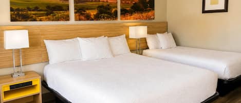A hotel room with two beds, a wooden headboard, bedside lamps, a small yellow nightstand, and a framed picture on the wall.
