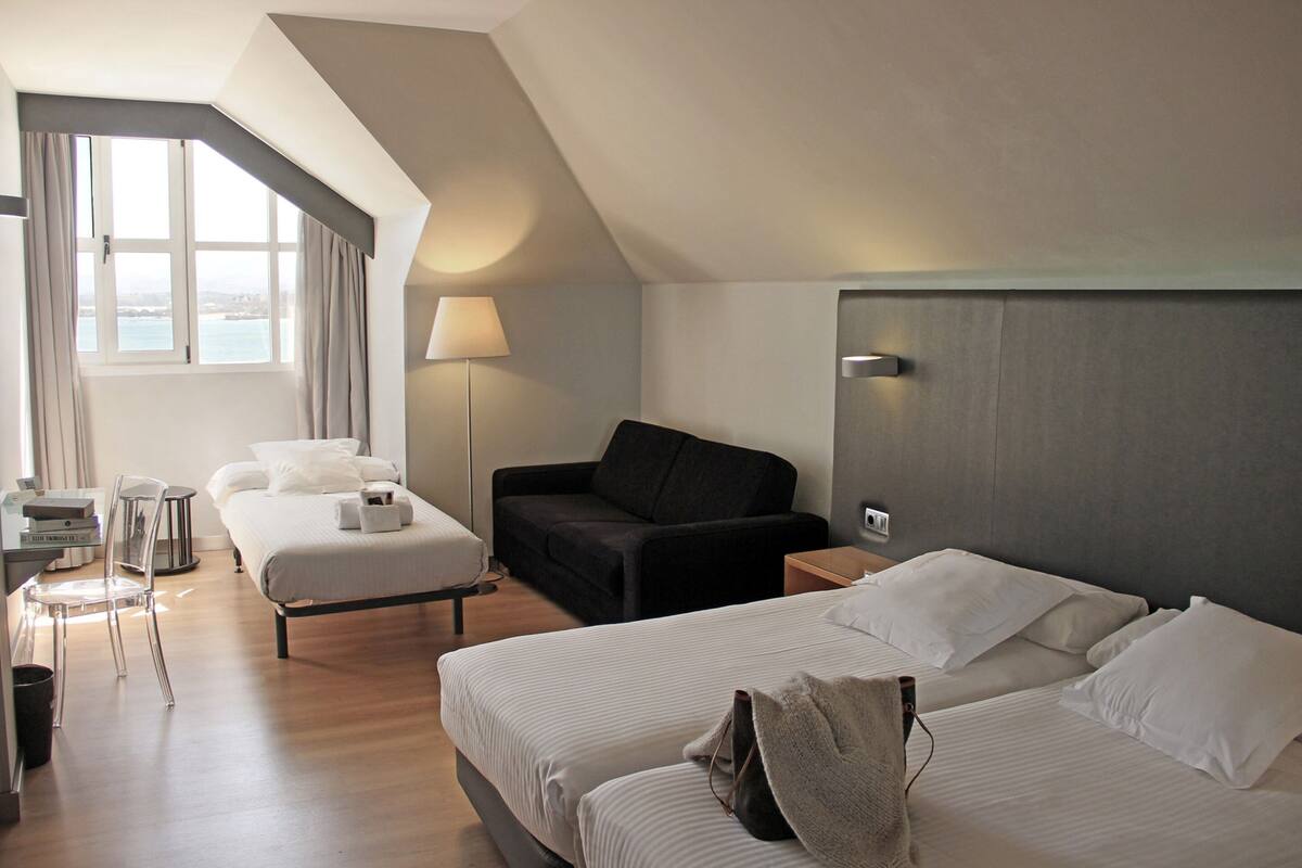 Habitación de hotel con dos camas, un sofá, un escritorio con silla, una lámpara y vista al mar.