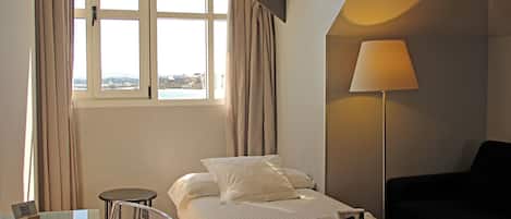 Habitación de hotel con cama, escritorio, silla, lámpara y vista de la ciudad a través de amplias ventanas.