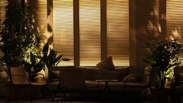 A cozy seating area with a sofa and chairs, surrounded by large windows with blinds, and potted plants.