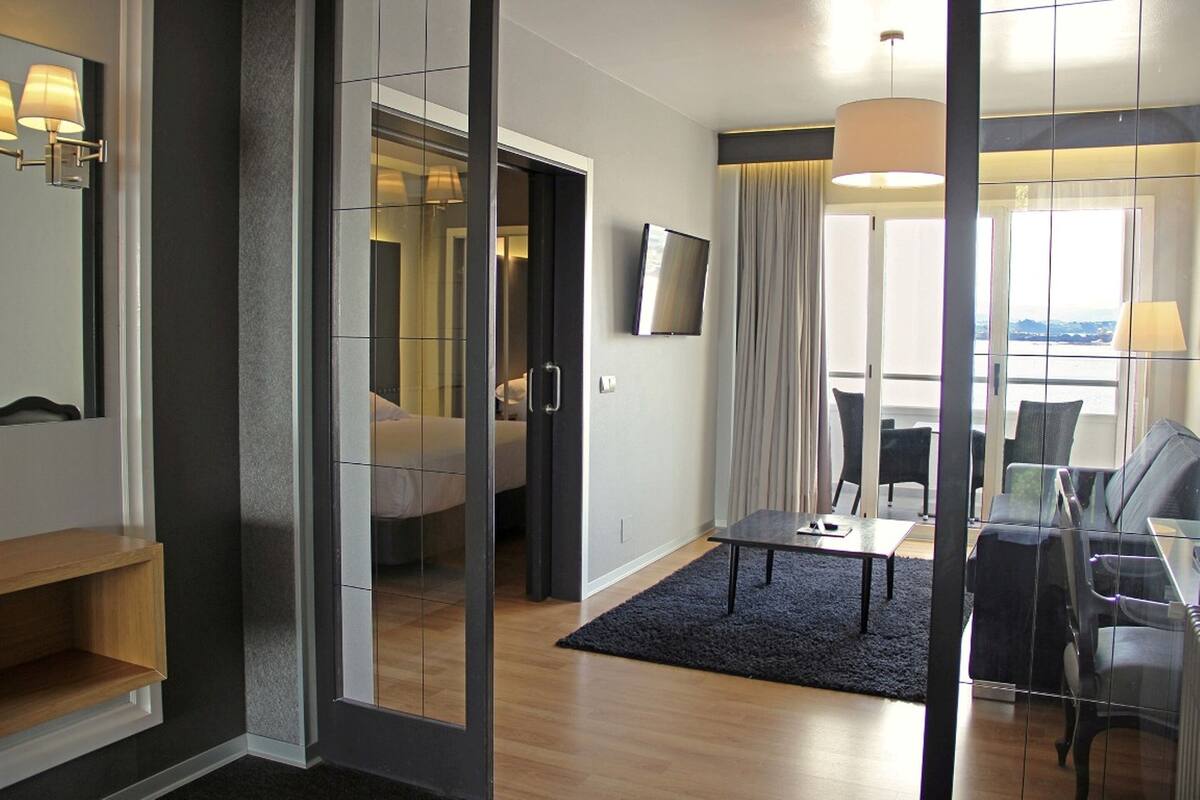 Una habitación de hotel moderna con una cama grande, un televisor, una zona de estar con una mesa y sillas, y vistas al exterior.