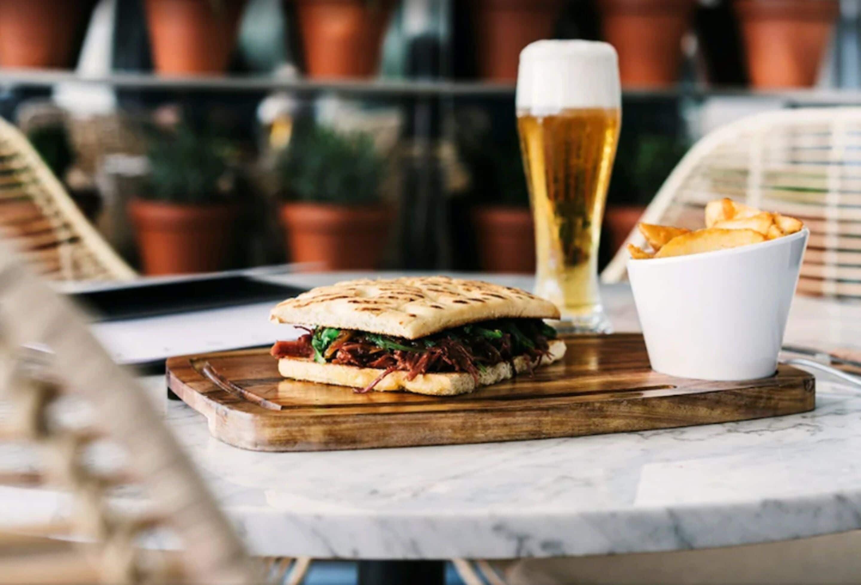 Un sándwich sobre una tabla de madera, una jarra de cerveza y un bol de papas fritas sobre una mesa.