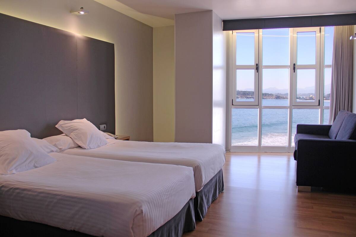 Habitación de hotel con dos camas, un sofá y un ventanal con vistas al mar.