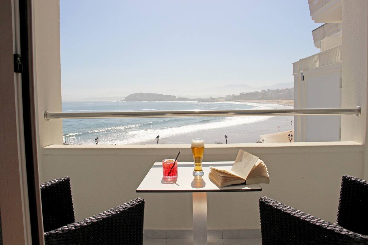 Un balcón con una mesa, una bebida y un libro, con vista a una playa donde hay gente y una montaña distante.