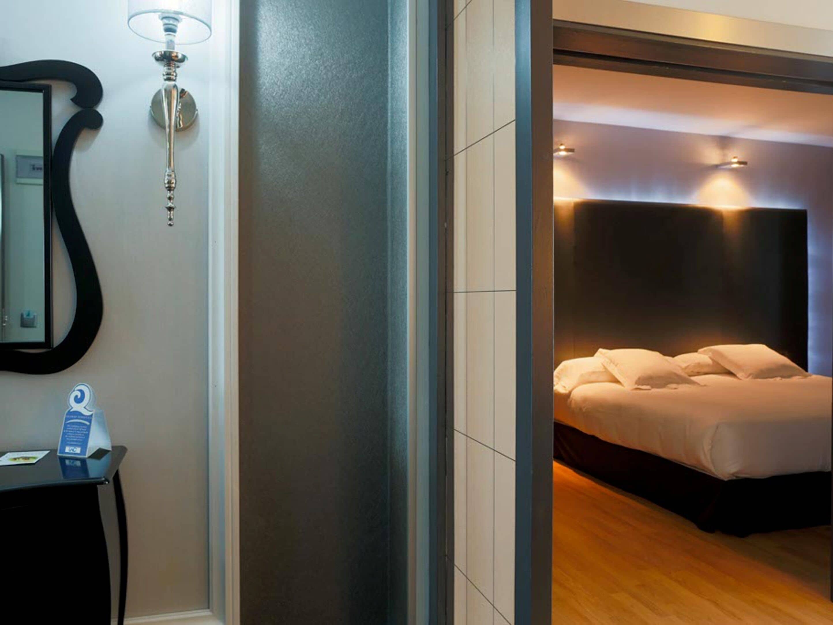 A modern hotel room with a large bed, a bedside table, a mirror, and a wall-mounted lamp.