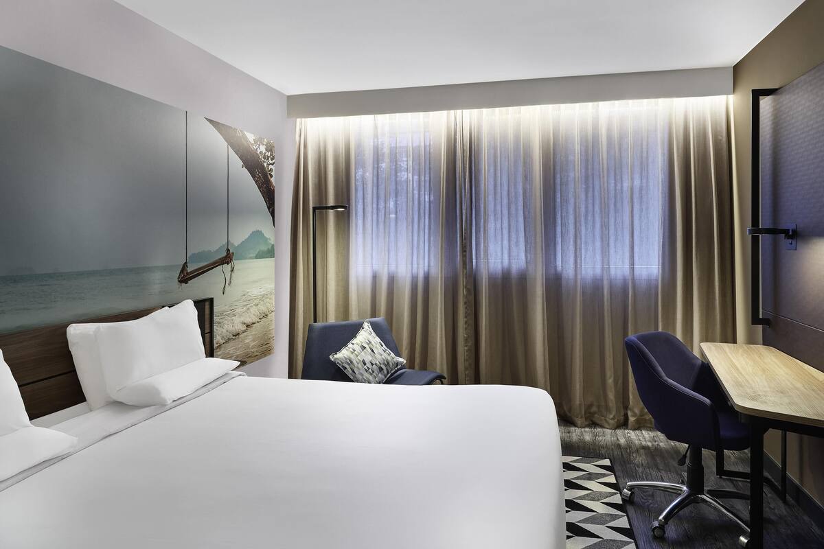 A hotel room with a bed, a desk, and a chair. There is a large window with curtains, a painting of a beach scene on the wall, and a view of the sea.