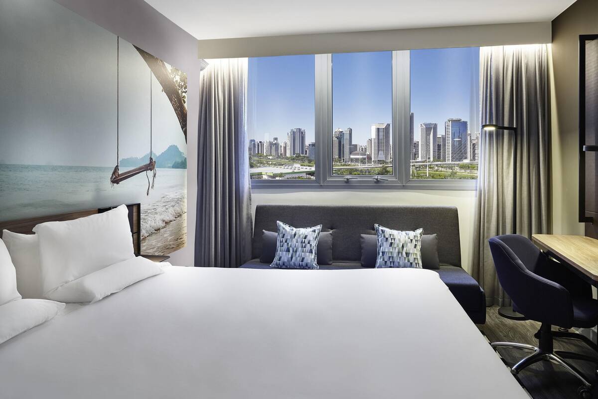 Een hotelkamer met een groot bed, een bureau en een uitzicht op een stadsbeeld door grote ramen.