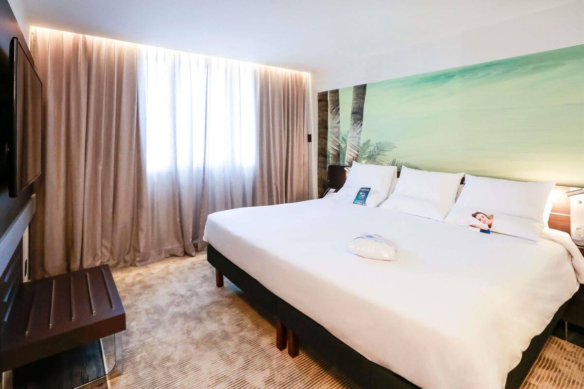 Een hotelkamer met een bed, gordijnen, een televisie en een schilderij aan de muur.