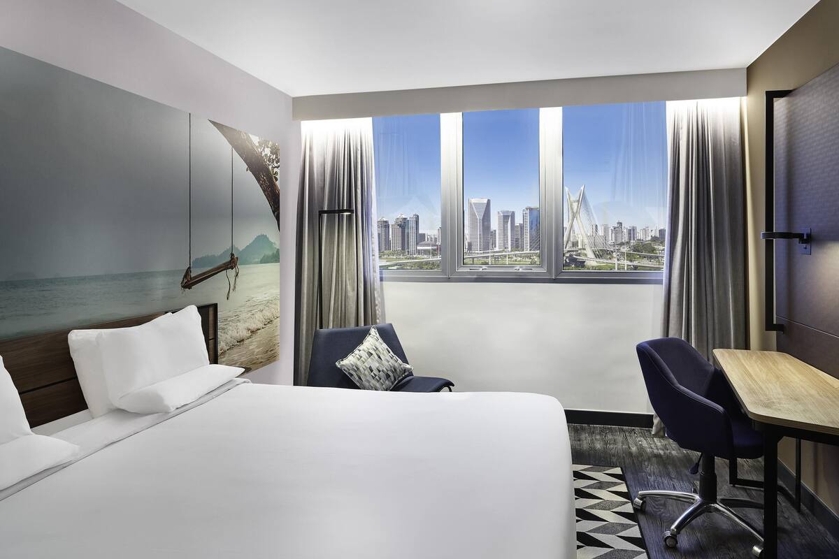 Een hotelkamer met een bed, een bureau, een stoel en een groot raam met uitzicht op de stad.
