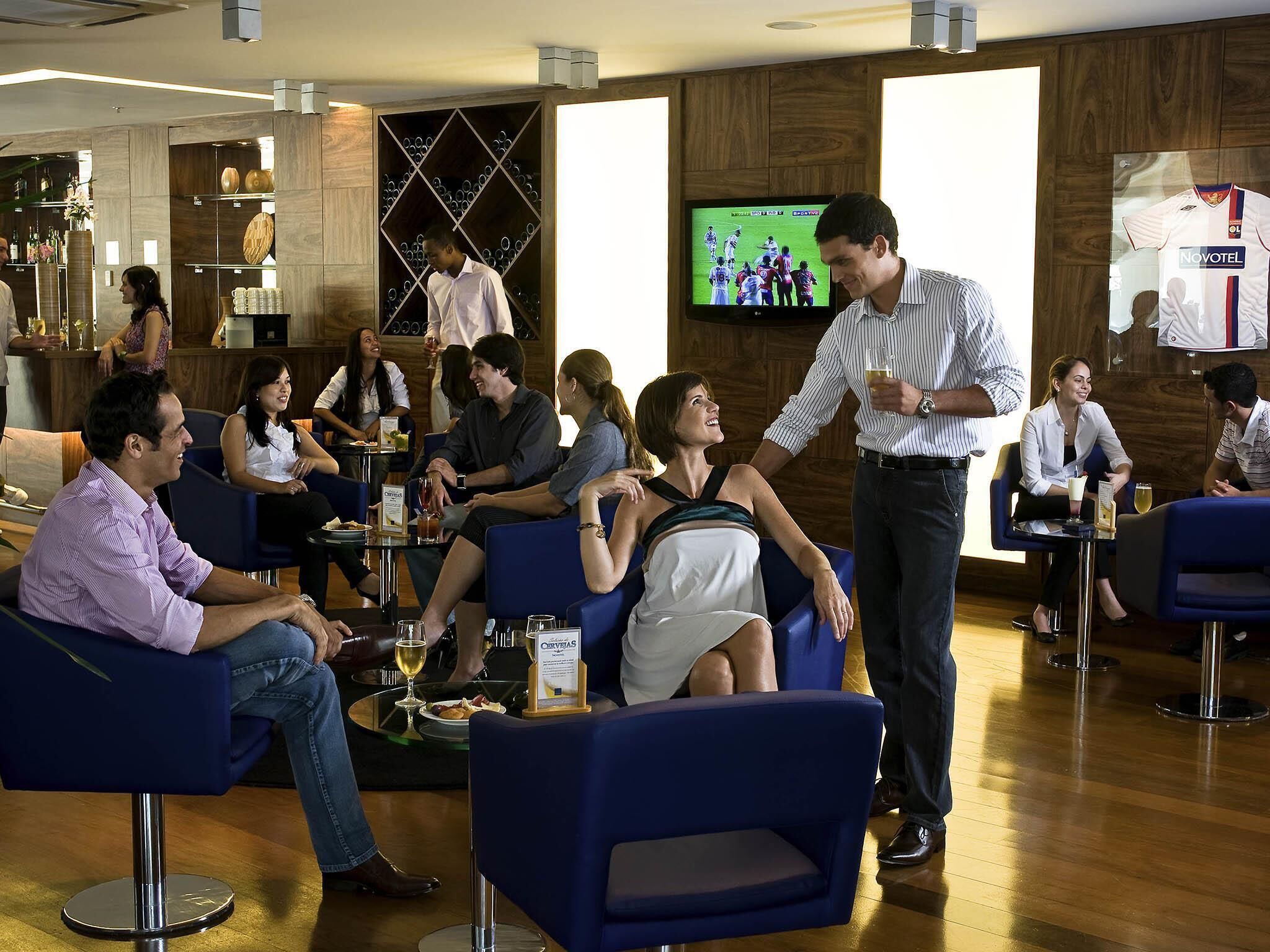 A social setting with people seated and standing, a television displaying a sports event, and a bar area with bottles and glasses.