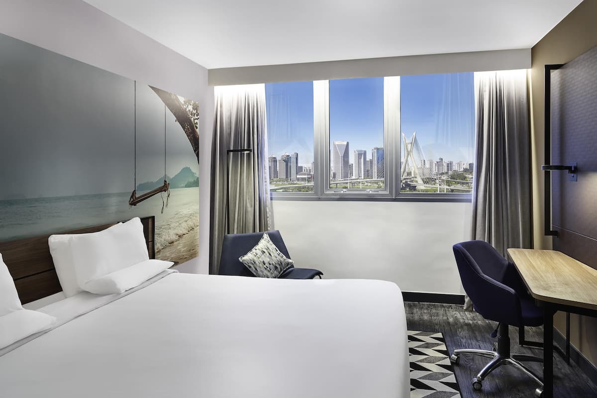 Habitación de hotel con cama, escritorio, silla y una ventana grande con vista a la ciudad.
