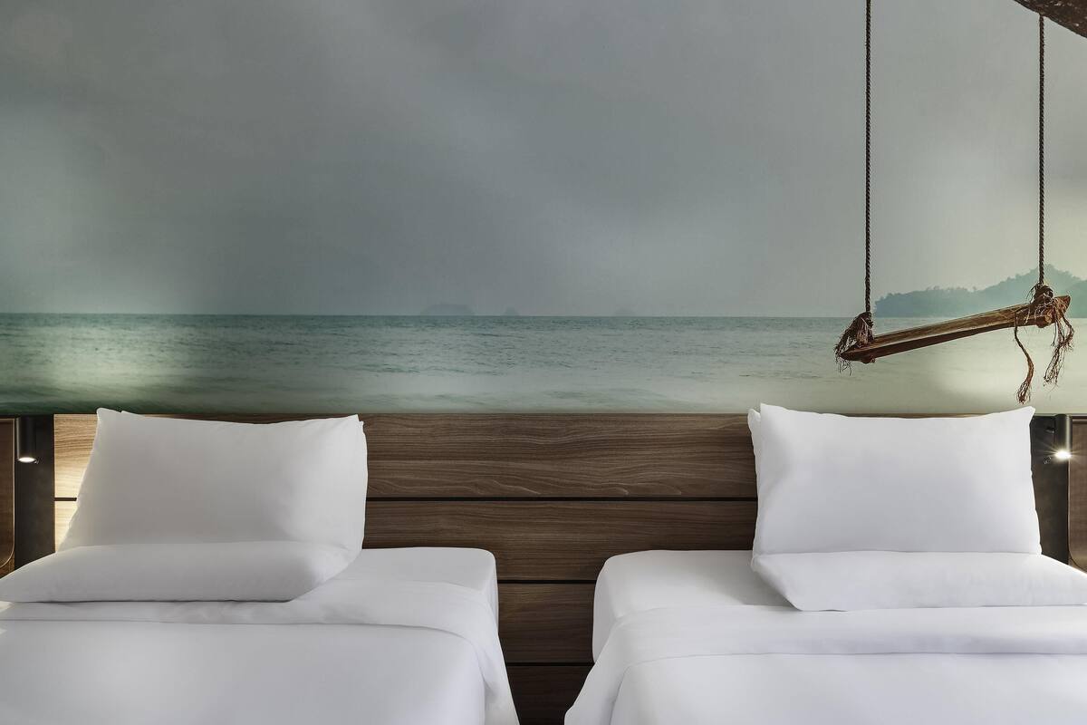 Een kamer met twee bedden, een houten hoofdbord en uitzicht op zee door een groot raam.