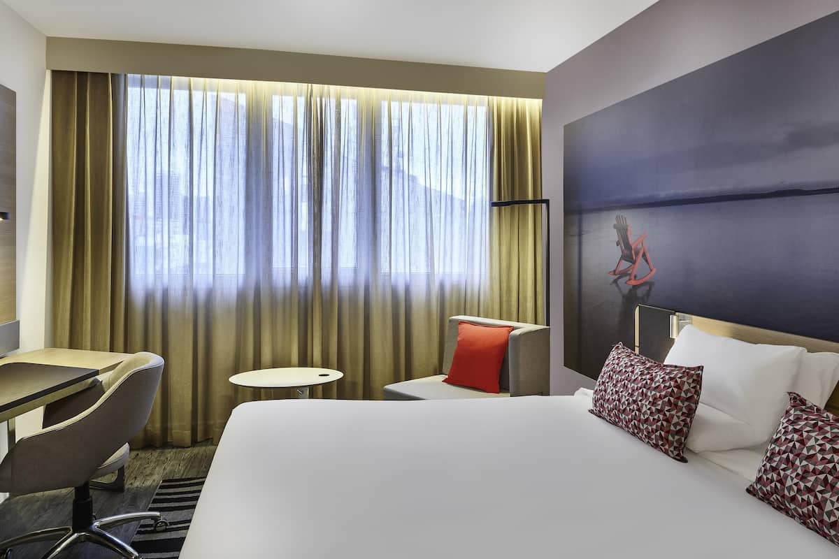 Habitación de hotel con cama, escritorio, silla, mesita pequeña y un amplio ventanal con cortinas.