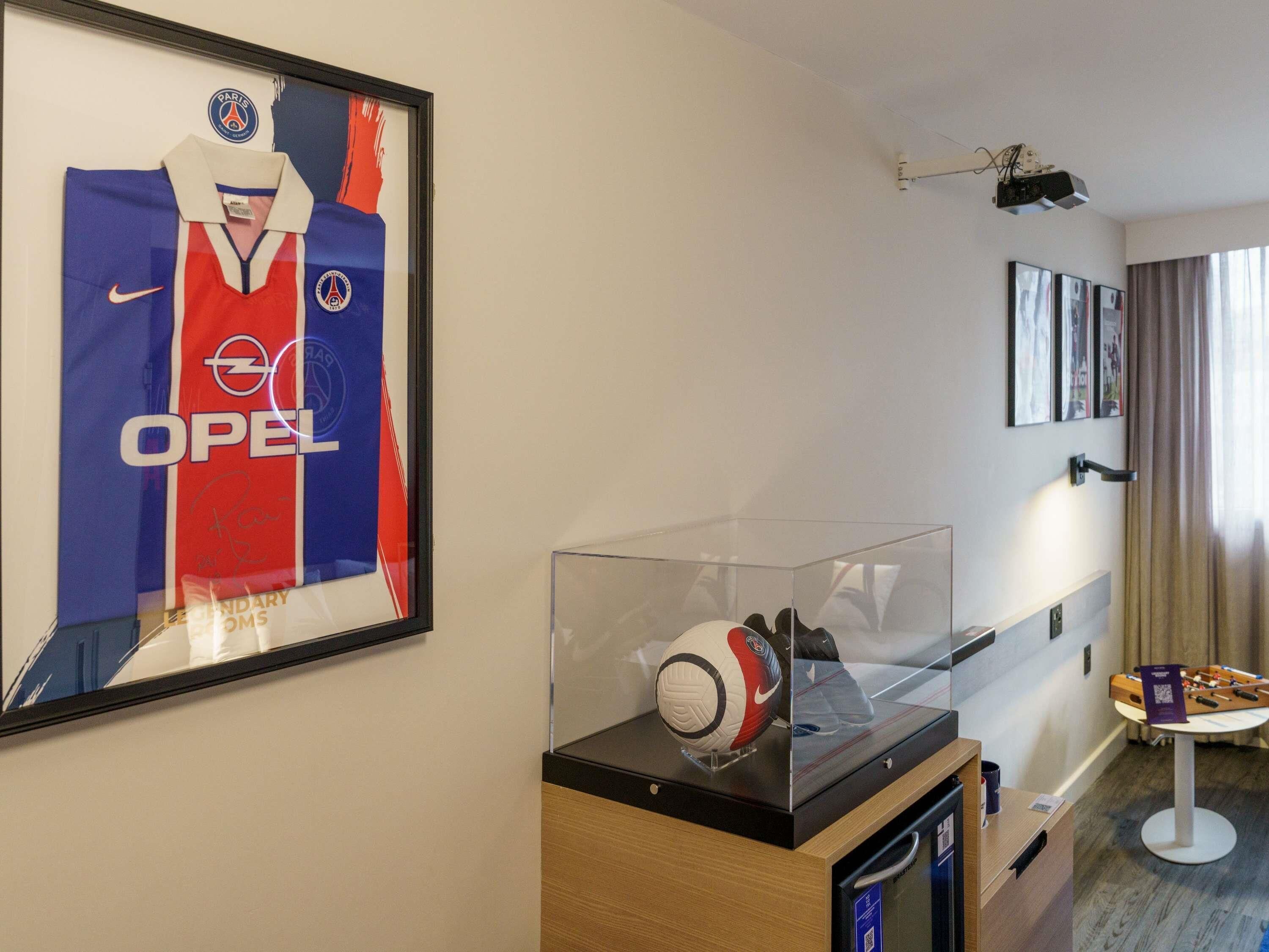 A hotel room with a framed Paris Saint-Germain jersey, a glass display case with a soccer ball, and a foosball table.