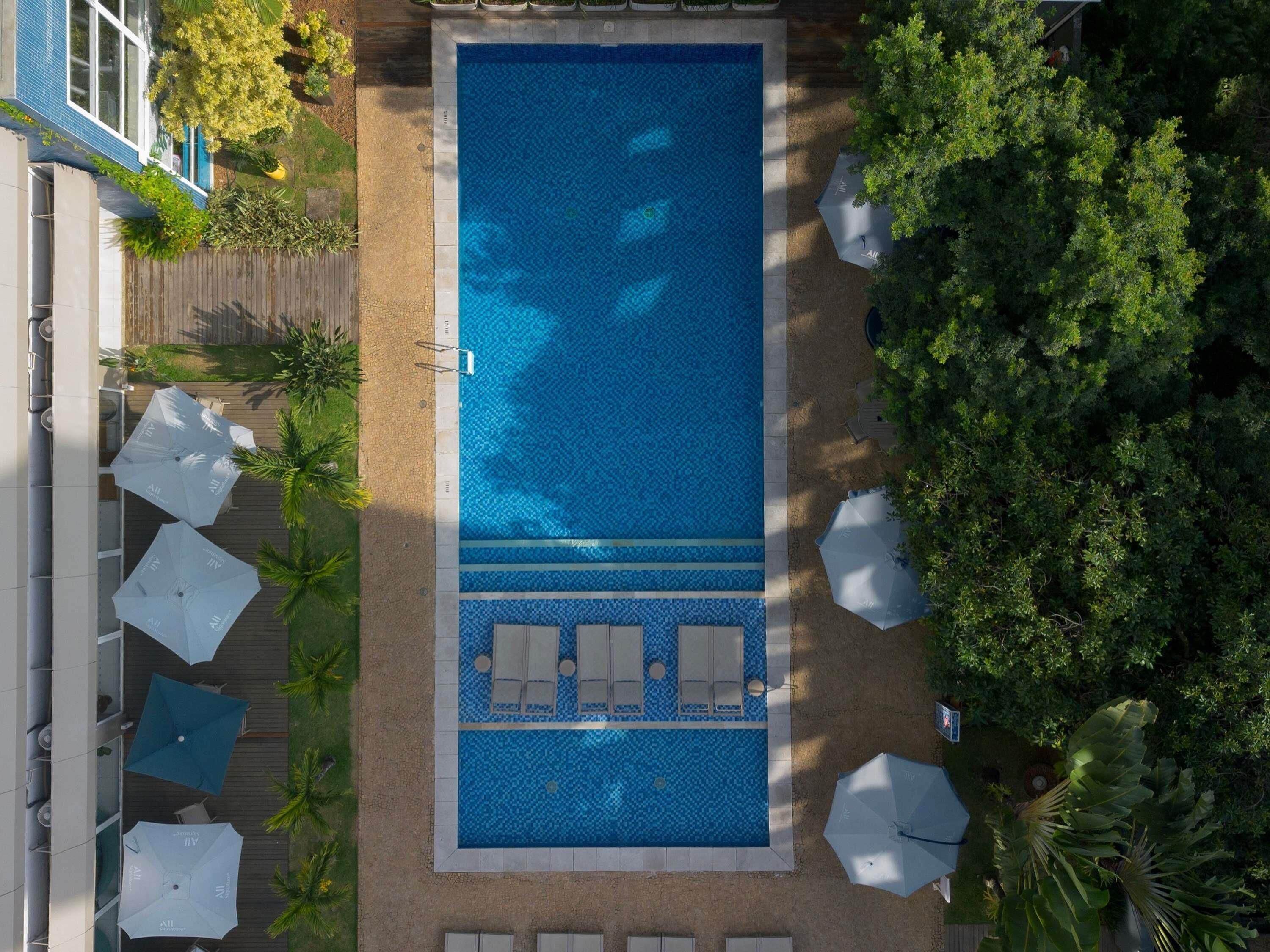 A rectangular pool with a tiled pattern, surrounded by lounge chairs and umbrellas, with trees and a building in the background.